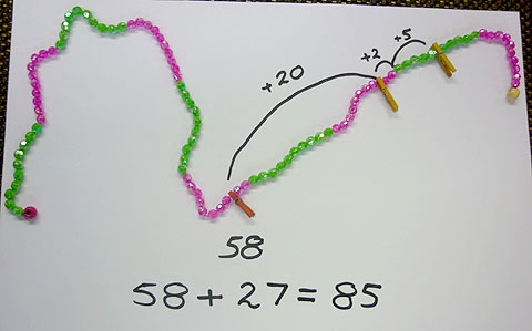 A string of beads, alternating every ten beads from green to pink. The problem is to add 27 to 58. First peg is at 58 with a jump of 20 to reach 78, then a jump of 2 to reach 80 then a jump of 5 to reach the answer of 85.
