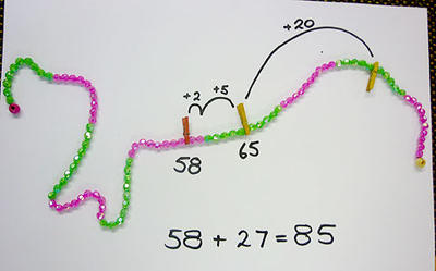 A string of beads, alternating every ten beads from green to pink. The problem is to add 27 to 58. First peg is at 58 with a jump of 2 to reach 60, then a jump of 5 to reach 65 then a jump of 20 to reach the answer of 85.